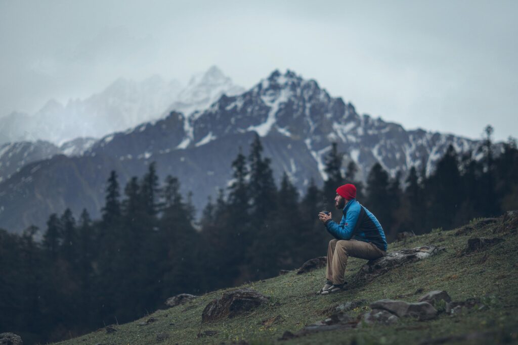 A man in colorful winter attire sits on a slope, enjoying the view of snow-capped Himalayan mountains in India.